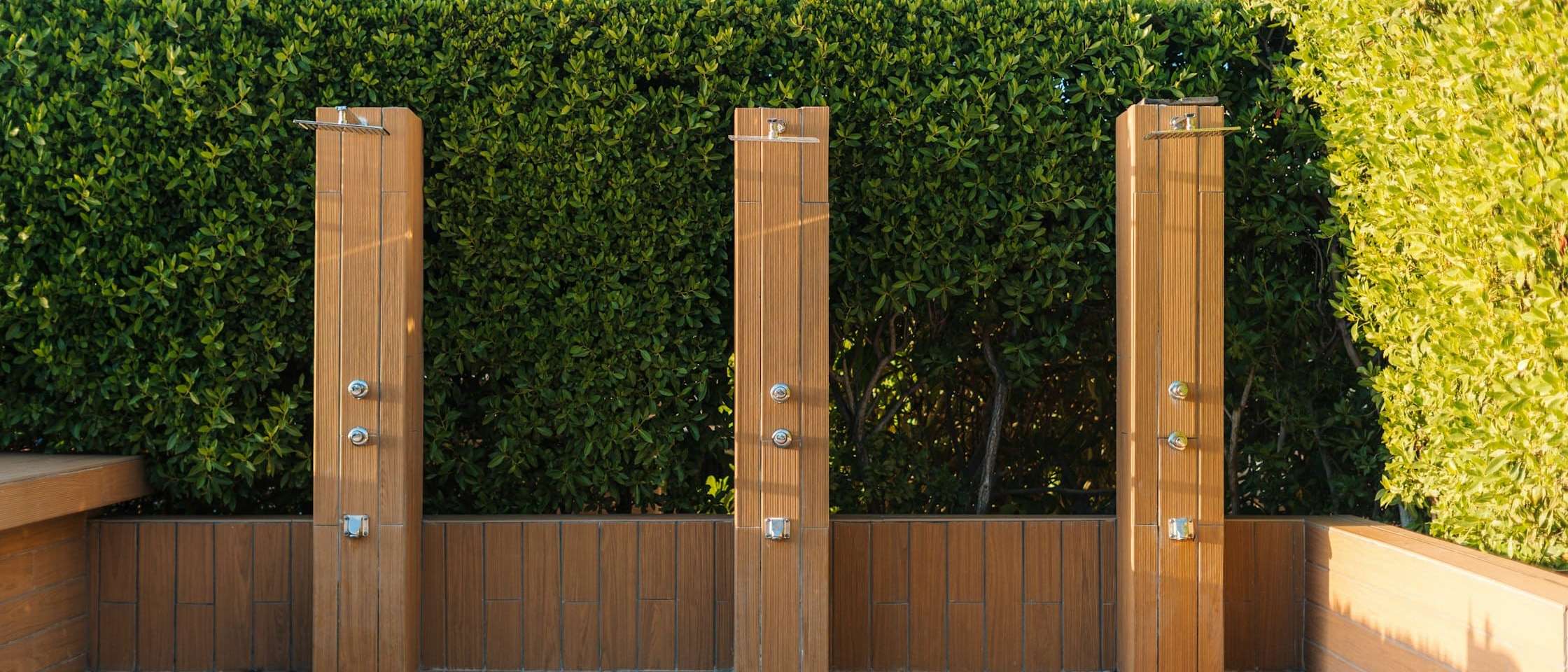 Tres duchas exteriores de madera sobre una tarima, delante de un seto