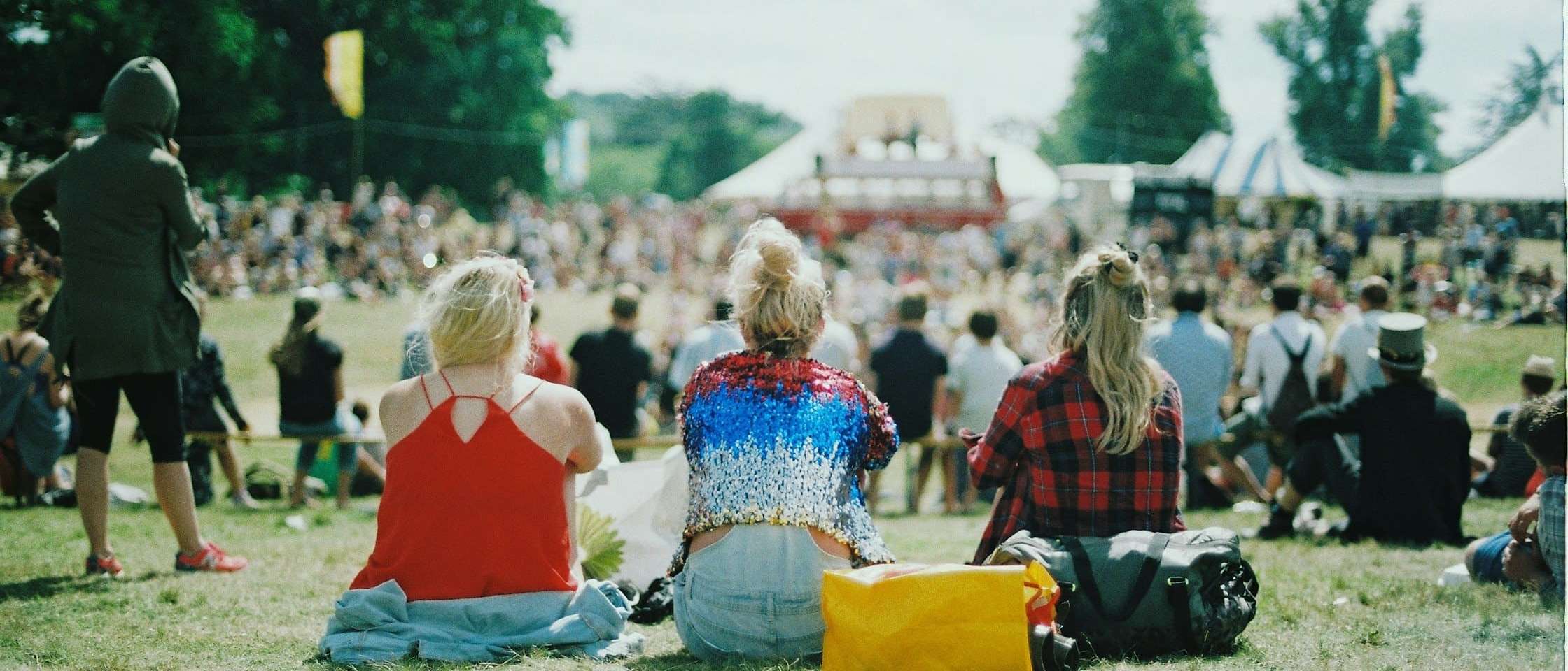 Público sentado en el césped de un festival de música de día, frente a las carpas